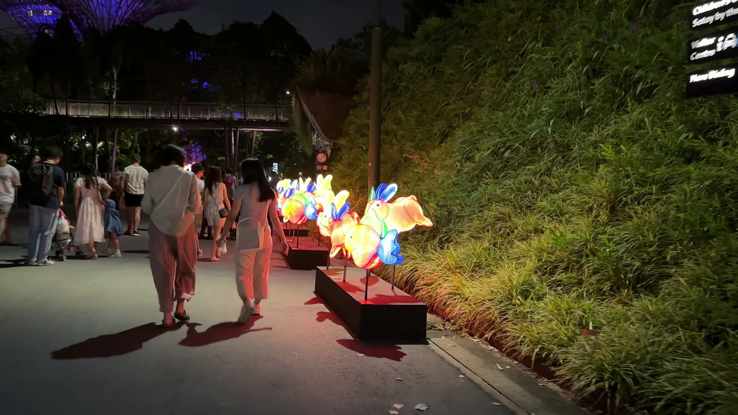 A nighttime scene with people walking along a pathway illuminated by colorful lanterns shaped like fish, surrounded by greenery